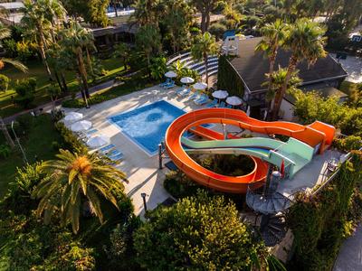 Una zona di parco acquatico con una grande piscina blu e uno scivolo d'acqua rosso in un ambiente tropicale.