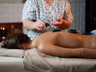 Eine Person liegt auf dem Bauch, während eine Masseurin eine Hot-Stone-Massage mit schwarzen Steinen durchführt.