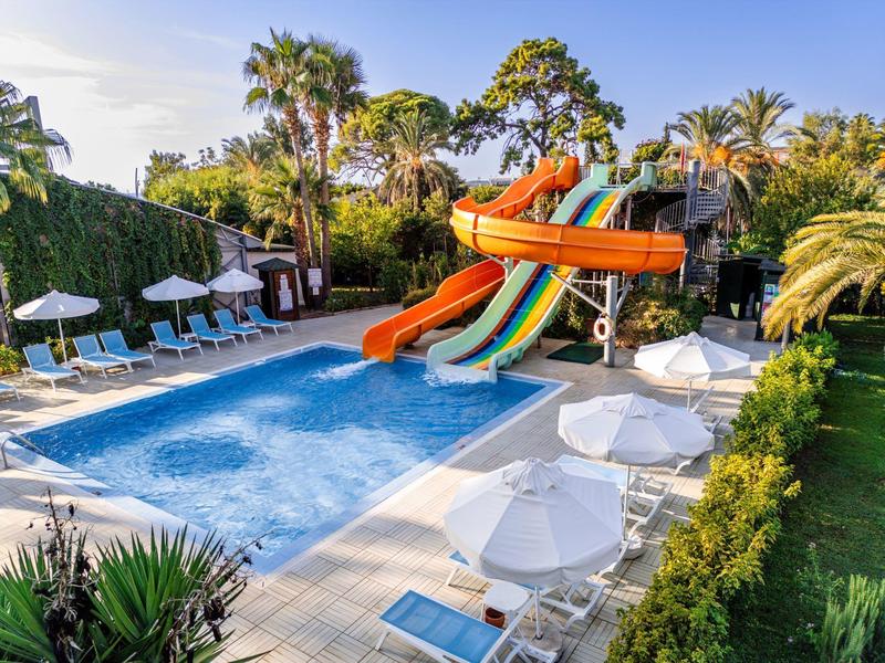 Außenpool mit bunten Wasserrutschen, weißen Schirmen und Palmen in sonniger Umgebung.
