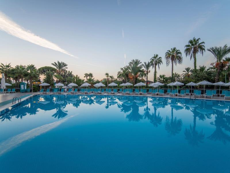 Großer, ruhiger Pool mit klar blauem Wasser, Palmen und Sonnenschirmen im Hintergrund bei Sonnenuntergang.