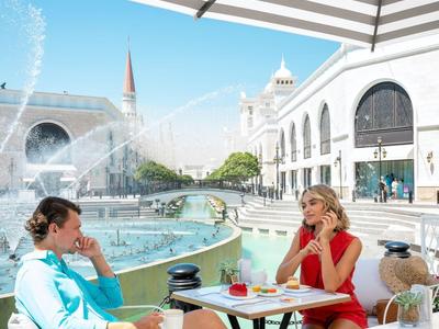 Pár odpočívá u venkovního stolu s fontánou a bílou budovou v pozadí.