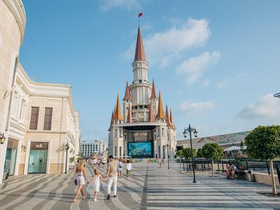 Lidé jdou po dlážděném náměstí s moderní budovou a zámkem v pozadí.