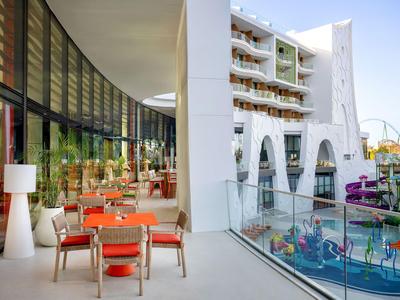 Moderne buitenruimte van een hotel met zitplaatsen en uitzicht op een attractiepark met ritten.