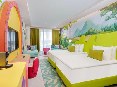 Modern hotelkamer met tweepersoonsbed, kleurrijk meubilair en een natuurmuurschildering.