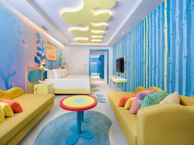 Kleurrijk ingerichte hotelkamer met gele banken, ronde tafels en bosachtige wanddecoraties.