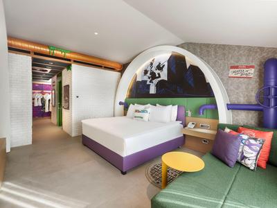 Modern hotelkamer met rond bed, kleurrijke muurschildering, groene bank en gele bijzettafels.