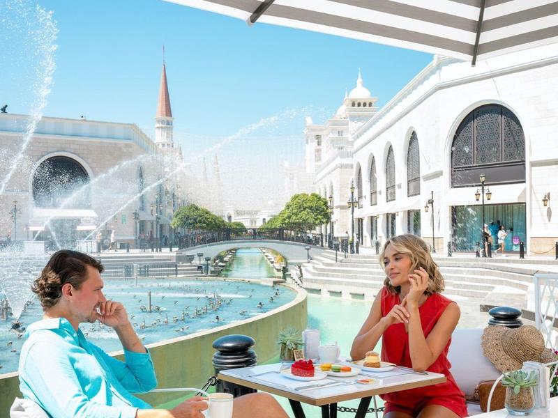 Twee mensen zitten buiten aan een tafel met drankjes naast een fontein bij zonnig weer.