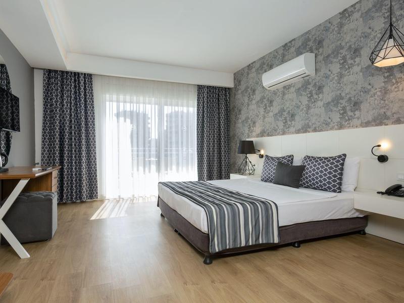 Chambre d'hôtel moderne avec grand lit, sol en bois et grande fenêtre lumineuse.