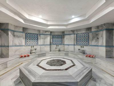 Großer türkischer Hamam mit zentralem Marmorbrunnen und Wandfliesen in Hellgrau und Blau.