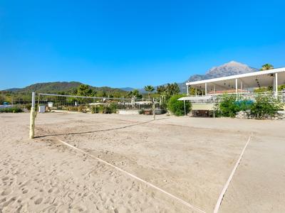 Un campo da beach volley sabbioso davanti a un edificio con vegetazione e montagne sullo sfondo sotto un cielo blu limpido.