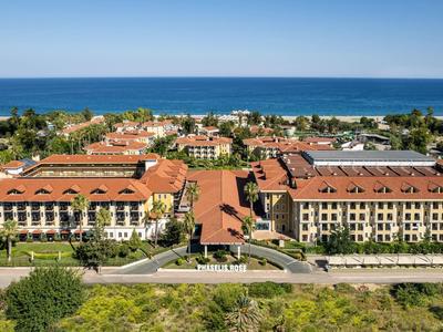 Großes, gelbes Hotel mit roten Dächern am Meer, umgeben von grünen Bäumen und blauem Himmel.