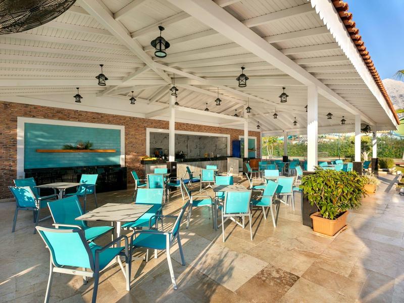 Außenbereich eines Restaurants mit türkisfarbenen Stühlen, Tischen, Pflanzen und Holzdecke.