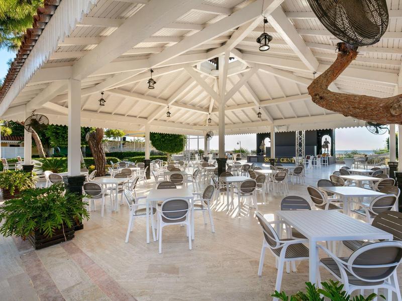 Überdachte Terrasse mit weißen Tischen und Stühlen, umgeben von Pflanzen und offenen Seiten.