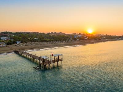 Sonnenuntergang über einem langen Steg am ruhigen Strand mit wenigen Häusern im Hintergrund.