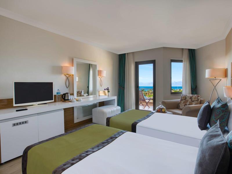 Hotel room with two beds, desk, TV, and sea view through window.