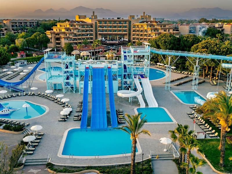 Water park with several water slides, pools, and sun loungers in front of a hotel building at sunset.
