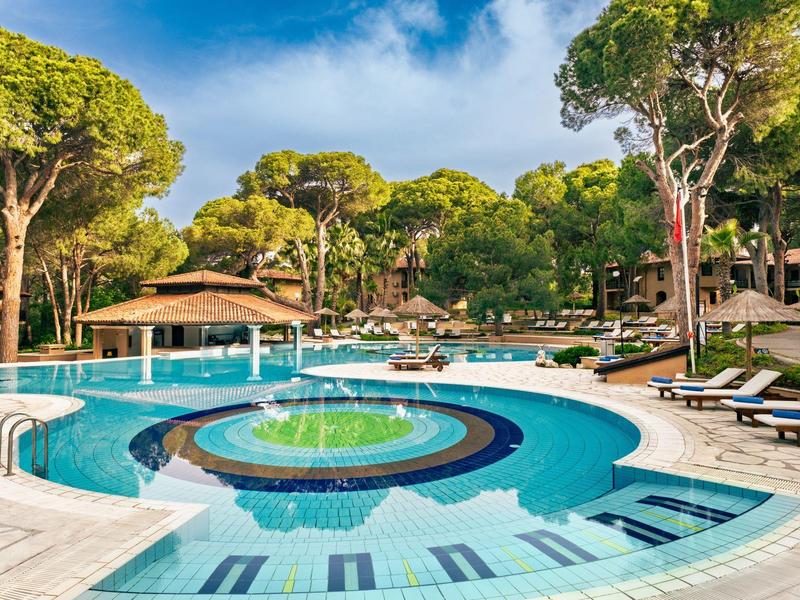 Round hotel pool with water fountain surrounded by sunbeds and pine trees under a blue sky.