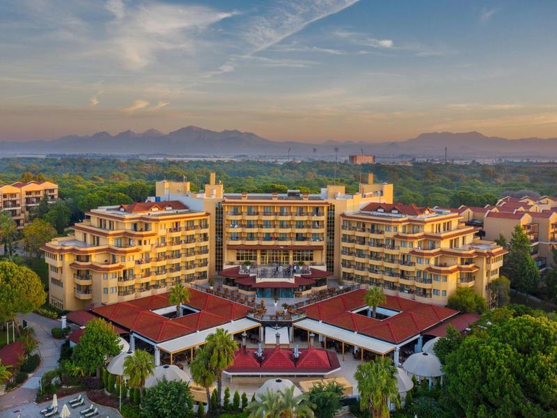 Large hotel resort with multiple buildings surrounded by trees and mountains in the background at sunset.