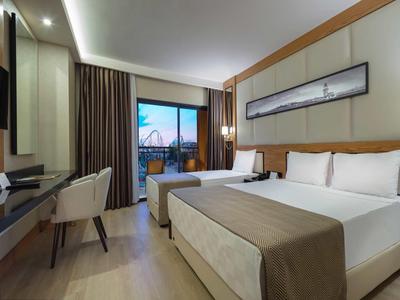 Chambre d'hôtel moderne avec lit double, TV, bureau et balcon avec vue sur la ville au coucher du soleil.