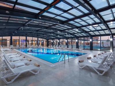 Jasny basen kryty z białymi leżakami pod nowoczesnym szklanym dachem.