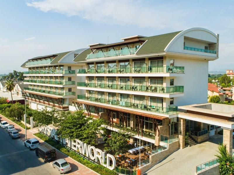 Großes modernes Hotelgebäude mit Balkonen, umgeben von Bäumen und geparkten Autos an der Straße.