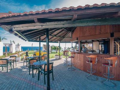 Offene Strandbar mit Holztheke und Sitzgelegenheiten auf gepflastertem Boden unter blauem Himmel.