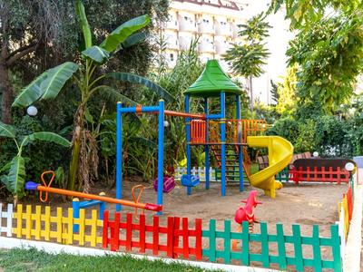 Parque infantil colorido con tobogán, estructura de escalada y zona de arena, rodeado de una valla multicolor y vegetación.