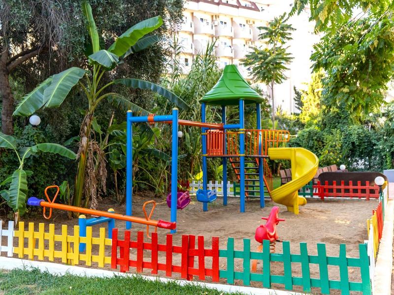 Parque infantil colorido con tobogán, estructura de escalada y zona de arena, rodeado de una valla multicolor y vegetación.