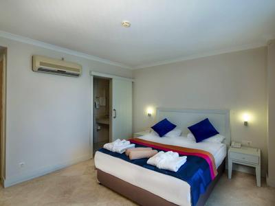 Habitación de hotel sencilla con cama doble, cojines azules, mesitas y baño al fondo.