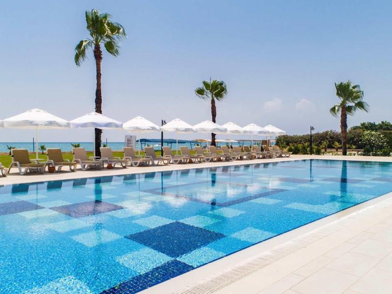 Piscina al aire libre con sombras blancas y palmeras junto al mar en un día soleado.