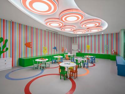 Sala infantil con patrón de rayas coloridas, mesas y sillas redondas y anillos luminosos en el techo.