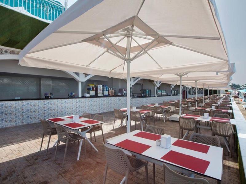 Área exterior de restaurante con mesas bajo grandes sombrillas junto a una piscina de hotel.