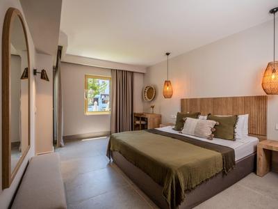 Hell eingerichtetes Hotelzimmer mit Doppelbett, großem Spiegel und Fenster mit Blick ins Freie.