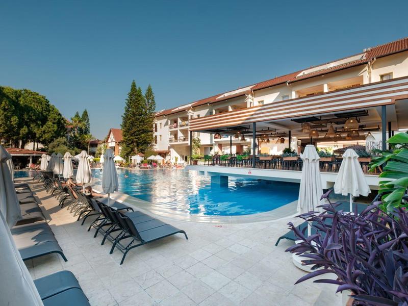 Hotelpool mit Liegestühlen und Sonnenschirmen neben einem großen Hotelgebäude bei klarem Himmel.