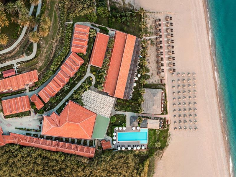 Luftaufnahme eines Hotels mit roten Dächern, Pool, Strandliegen und blauem Meer am Sandstrand.