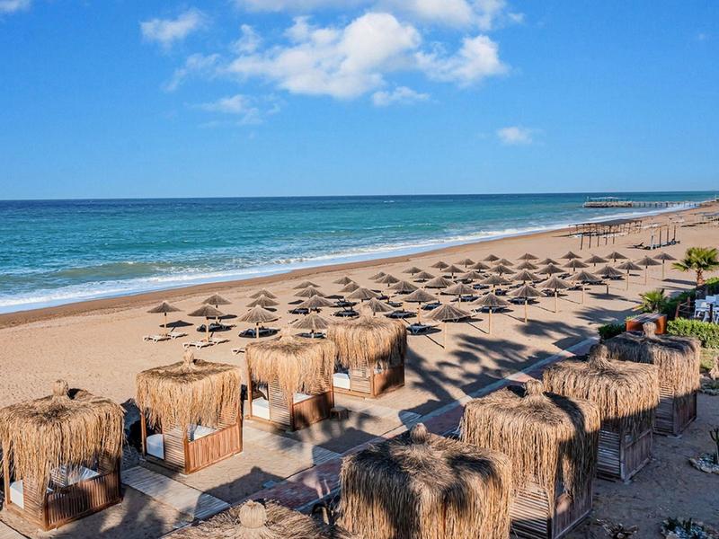 Weitläufiger Sandstrand mit Reihen von Strohdach-Sonnenschirmen und blauer Meereslinie unter blauem Himmel.