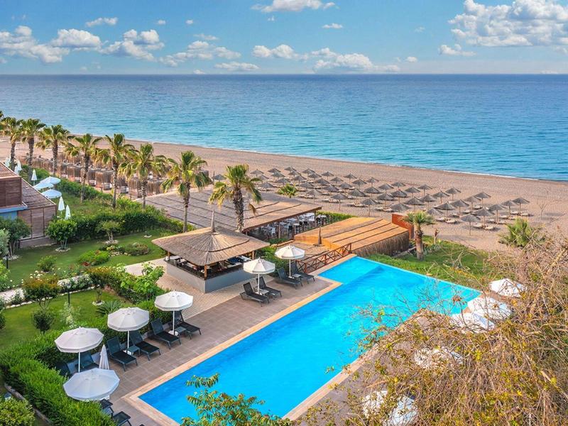Ein Swimmingpool mit blauer Wasserfläche neben einem Sandstrand und Palmen unter blauem Himmel.