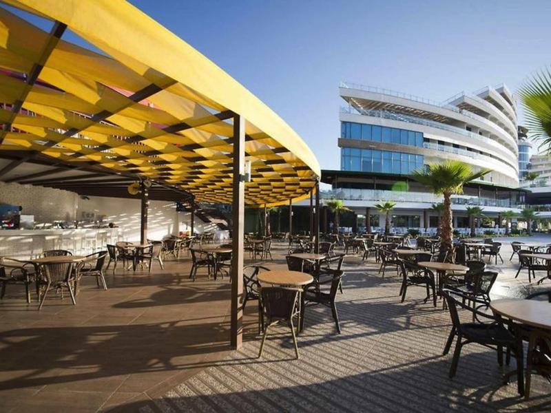 Große Terrasse mit Tischen und Stühlen, teilüberdacht durch gelbes Dach, modernes Gebäude im Hintergrund.