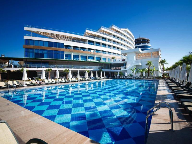 Moderner Hotelbau mit Glasfassade, großem blauen Pool und Sonnenschirmen unter klarem Himmel.