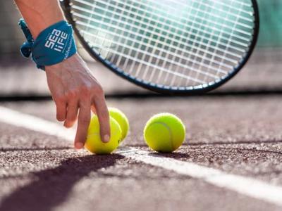 Ruka s náramkovými hodinkami pokládá tři tenisové míčky na tenisový kurt s raketou v pozadí.