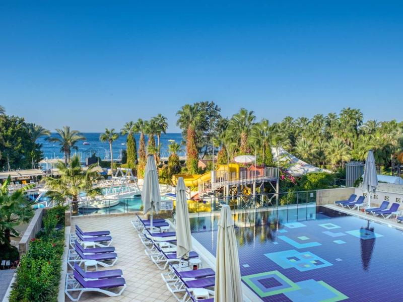 Großer Pool mit Sonnenschirmen, Liegen und Palmen vor blauem Himmel am Meer.