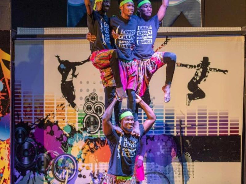 Drei Tänzer in bunter Kleidung bilden eine Pyramide auf einer beleuchteten Bühne mit Graffiti-Hintergrund.