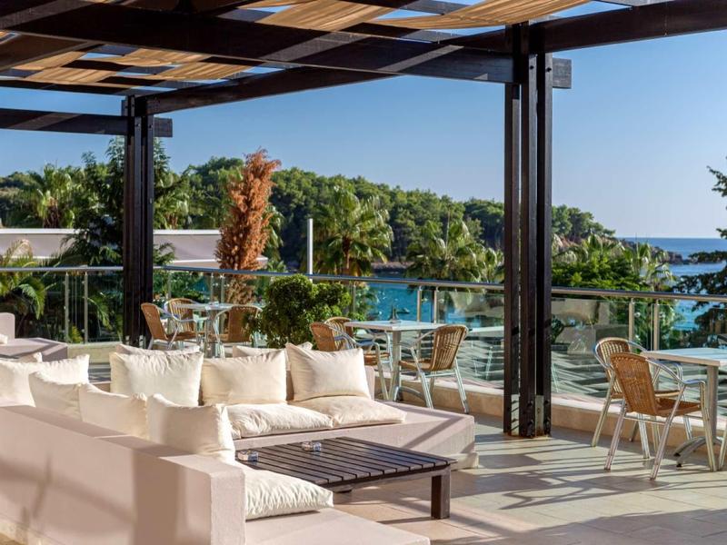 Terrasse mit weißen Sofas, Tischen, Stühlen und Blick auf grünen Garten und Meer im Hintergrund.