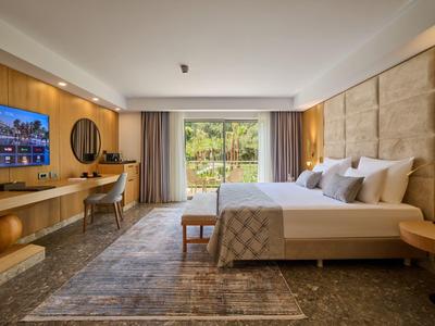 Modernes Hotelzimmer mit großem Doppelbett, TV, Schreibtisch und großer Fensterfront zum Garten.