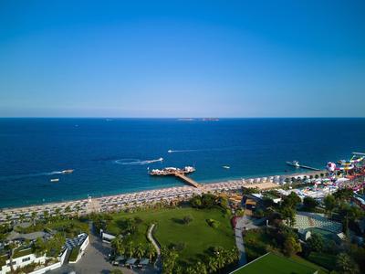 Strand mit Liegestühlen, Pier und blauem Meer, daneben Grünflächen und Häuser unter klarem Himmel.
