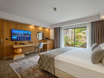 Modernes Hotelzimmer mit großem Bett, Schreibtisch, TV und Zugang zu einem Balkon mit Gartenblick.