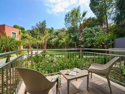 Balkon mit zwei Stühlen und Tisch, umgeben von grünen Pflanzen und Bäumen, unter blauem Himmel.