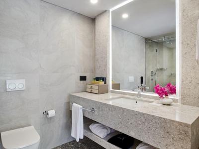 Moderner Bad mit grauen Fliesen, großem Spiegel und beleuchtetem Waschtisch mit Handtüchern und Blumen.