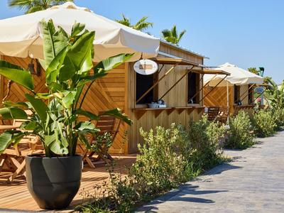Holzhütten mit Sonnenschirmen, grünen Pflanzen und Holzstühlen neben einem Gehweg bei blauem Himmel.