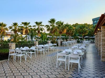 Outdoor-Terrasse mit weißen Stühlen und Tischen auf gemustertem Boden, Palmen im Hintergrund.